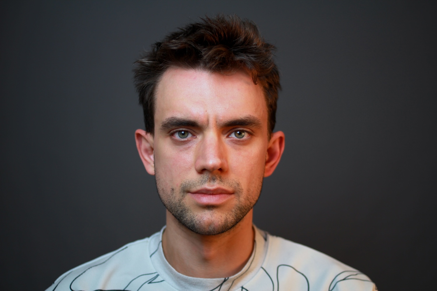 Portrait picture of Serafin Dinges. White masculine figure with brown hair, green eyes and a stubble wearing an off-white t-shirt with lines on it.